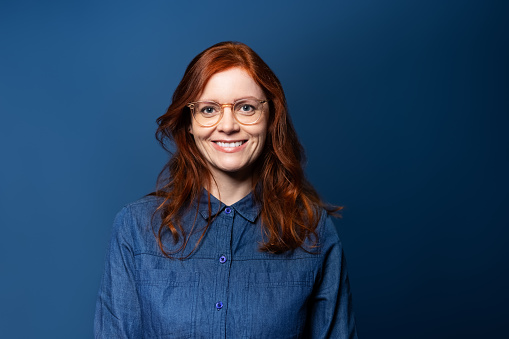 Smiling mature woman with red hair wearing eyeglasses looking at camera. Woman in denim shirt standing on blue background.