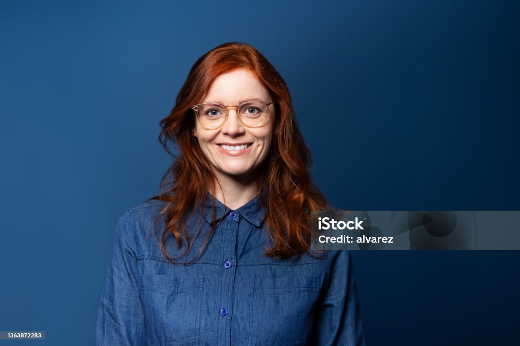 Portrait of a smiling mature woman with red hair on blue studio background Smiling mature woman with red hair wearing eyeglasses looking at camera. Woman in denim shirt standing on blue background. Portrait Stock Photo