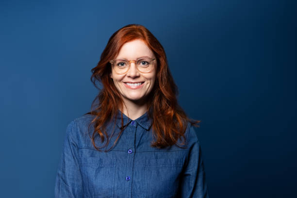 Portrait of a smiling mature woman with red hair on blue studio background Smiling mature woman with red hair wearing eyeglasses looking at camera. Woman in denim shirt standing on blue background. eyewear photos stock pictures, royalty-free photos & images
