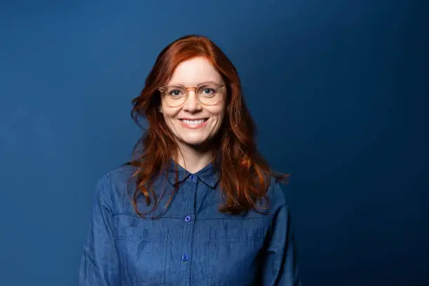 Smiling mature woman with red hair wearing eyeglasses looking at camera. Woman in denim shirt standing on blue background.