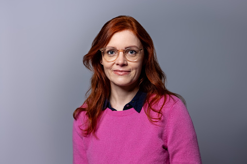 Beautiful mature woman with red hair being photographed in a studio. Female in casuals and eyeglasses looking at camera against gray background.