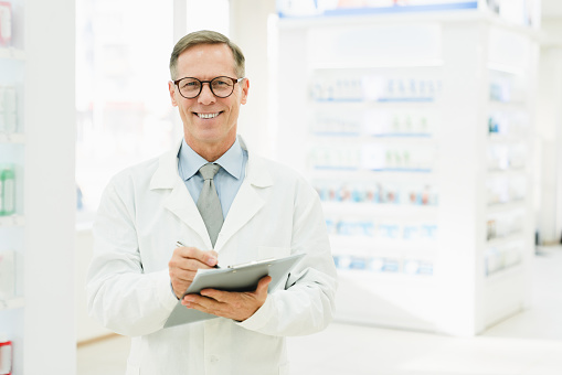Successful male mature chemist pharmacist druggist in white medical coat looking at camera writing on clipboard side effects, active substance, prescriptions standing at pharmacy drugstore
