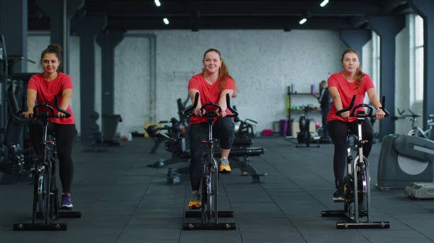 grupo de meninas atléticas realizando exercícios de treinamento de equitação aeróbica em bicicleta estacionária de ciclismo em academia - pedal bicycle sports training cycling - fotografias e filmes do acervo