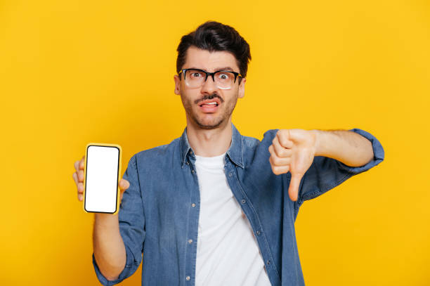 istock Caucasian handsome stylishly dressed guy with negative facial expression holds smartphone with blank white screen for your text, shows thumb down, stands on isolated orange background, looks at camera 1363884040