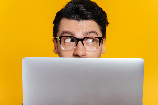 Amazed excited caucasian young adult guy with glasses peeking out from behind laptop, looking surprised to the side while standing against isolated orange background, close-up