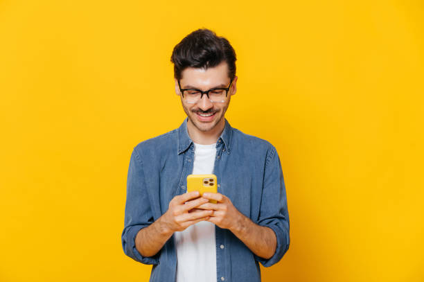 istock Stylish handsome positive caucasian guy is using his smartphone, texting with friends, browsing the Internet and social networks, standing on an isolated orange background, smiling 1363884043