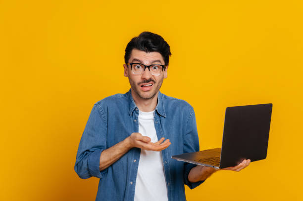 Discouraged displeased unshaven caucasian guy in glasses and in denim shirt, holds a laptop, looks confused at the camera, holding his head, without mood, stands on an isolated orange background stock photo