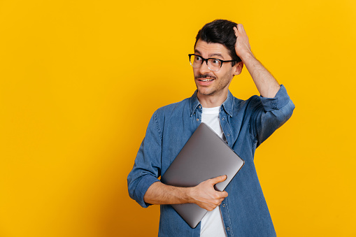 Surprised puzzled discouraged guy in glasses, in a denim shirt, holds a laptop at hand, looks thoughtfully towards empty space aside touching his head, stands on isolated orange background, copy space