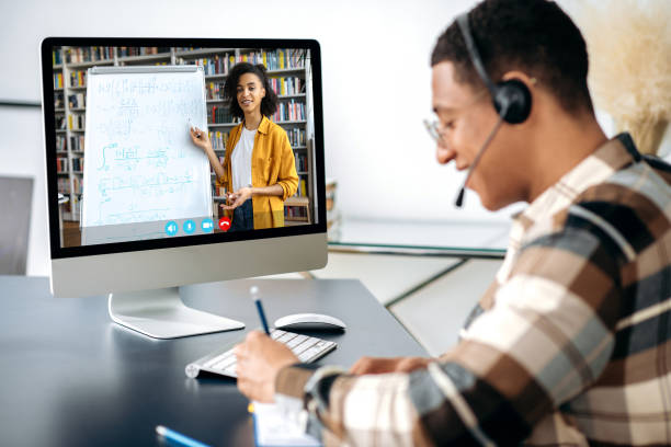 Distant learning, webinar, online lecture. Hispanic clever male student, sits at work desk, uses computer, listening to an online lecture, taking notes, female african american teacher on the screen