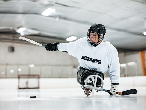 Mature Disabled Latin woman practising sledge hockey. She is a double leg amputee and her left hand is disfigured with no fingers. She is wearing full sledge hockey gear with jersey featuring her own print with universal symbols of various disabilities. She is also wearing full protection hockey helmet with face mask. Interior of hockey rink.