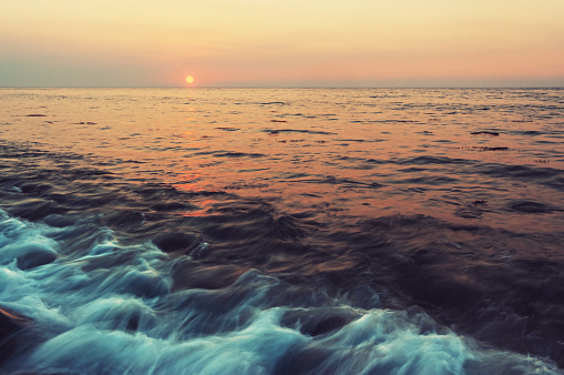 This image captures the serene beauty of the Karachi ocean at sunset. The golden hues of the setting sun paint a tranquil scene where waves gently lap against prominent rocks emerging from the shallow waters.