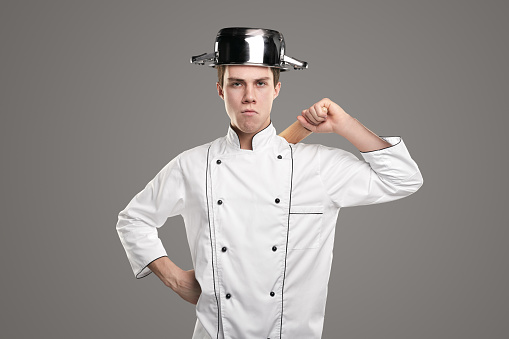 Confident male chef in white uniform and saucepan on head ready to war looking at camera on gray background