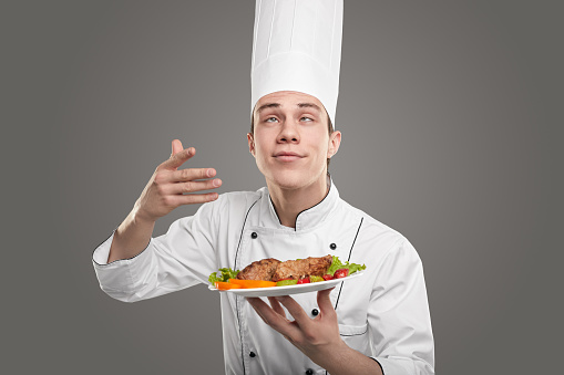 Funny chef in uniform standing with plate of meat with vegetables and smelling aromatic flavor, while closing eyes from pleasure in studio against gray background