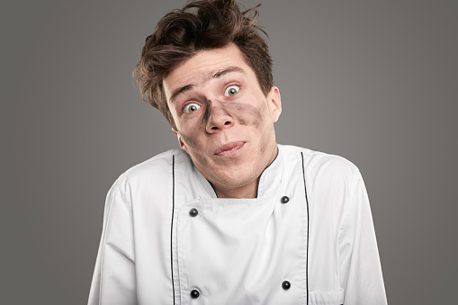 Funny young cook with soot on face shrugging shoulders and looking at camera while apologizing for mistake against gray background
