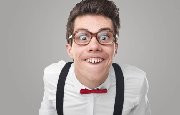Comic male nerd in eyeglasses and formal wear grimacing and showing teeth while looking at camera on gray background