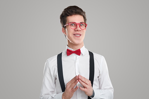 Crafty young man in red glasses looking at camera with tricky smile and making steeple gesture while scheming against gray background