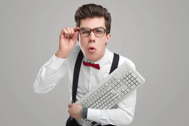 Goofy young man with computer keyboard looking and camera and adjusting glasses while working against gray background