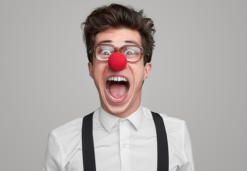 Funny male nerd in glasses and red clown nose yelling on gray background in studio and looking at camera