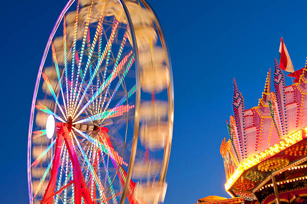 Carnival Ferris Wheel Lights Blur With Motion At Night The colored lights on a ferris wheel blur against a deep blue sky at a county fair at dusk. midway fair stock pictures, royalty-free photos & images