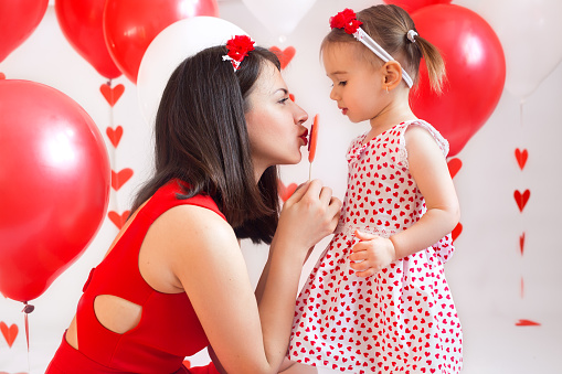 Mother and daughter with heart-shaped lollipops on a white background with decor for Valentine's Day. The woman's red dress and the daughter's white heart-shaped dress