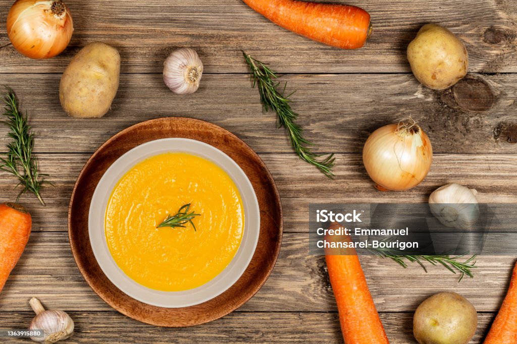 Carrot and potato soup bowl on drift wood table with a spread of vegetables, garlic, and rosemary. Flat lay. Carrot and potato soup in a beige bowl on a drift wood table with a spread of onions, potatoes, carrot, garlic and rosemary. Flat Lay, top view. Appetizer Stock Photo