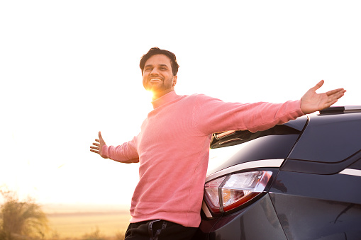 Happy young man leaning on car with arms outstretched by field