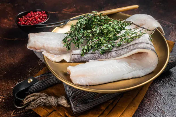 Whitefish fillet, raw Haddock fish on plate with rosemary and thyme. Dark background. Top view.