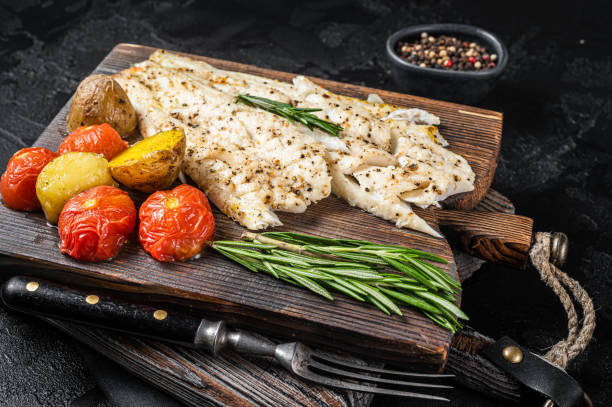 filé de peixe haddock assado na tábua de madeira com tomate e batata. fundo preto. vista superior - cod fillet fried prepared fish - fotografias e filmes do acervo