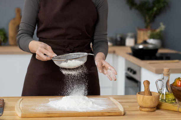 femme contemporaine en tablier tamisant la farine près de la table de cuisine - sifting photos et images de collection