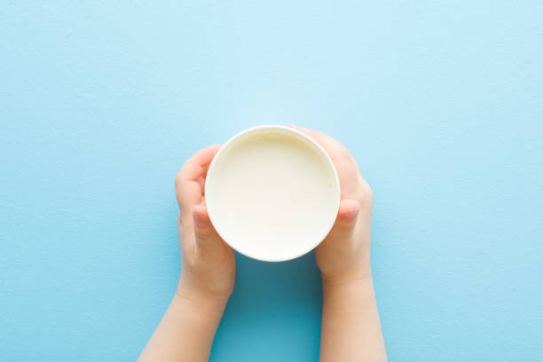 Toddler hands holding white plastic glass of fresh milk on light blue table background. Pastel color. Closeup. Point of view shot. Daily healthy drink. Top down view. stock photo