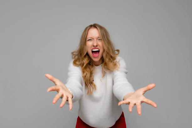 giovane donna irritata che urla disperatamente con la bocca spalancata, guardando la telecamera con espressione arrabbiata, indossa un maglione bianco, in piedi su sfondo grigio - mouth open women displeased anger foto e immagini stock