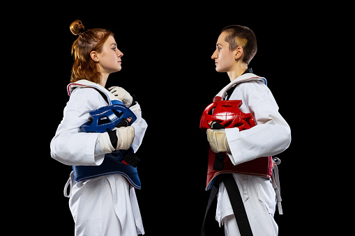 Half-length portrait of two young girls, taekwondo athletes posing isolated over dark background. Concept of sport, education, skills, workout, health. Sportsmen wearing white doboks
