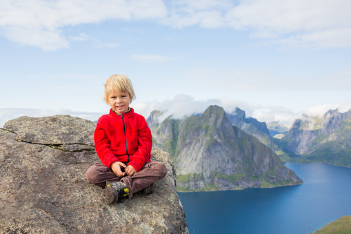 Cute child, sitting on top of the mountains and looking down on Reine after climbing Reinebringen treeking path with lots of stairs, Lofoten, northern Norway