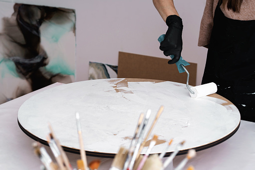 The artist works with a roller in her hand, applying paint to a round canvas in her workshop. Young artist paints in the home office.