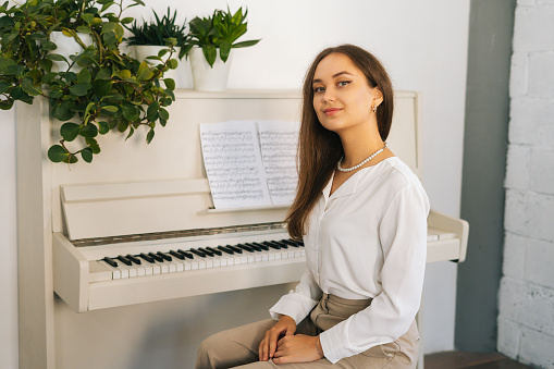 Portrait of smiling young woman pianist looking at camera sitting at classical white piano in classroom. Successful attractive female musician performer preparing for classes at home studio.