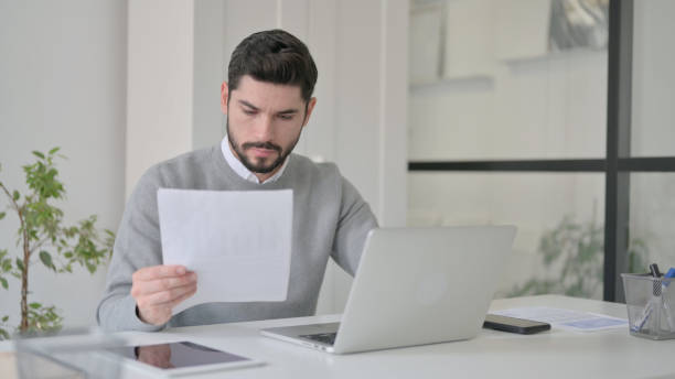 istock Young Man with Laptop Reading Documents in Office 1363974387