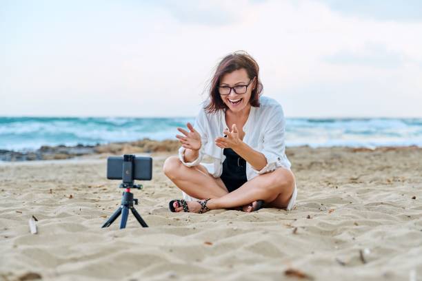 istock Middle aged woman sitting on beach with smartphone using video call 1363978132