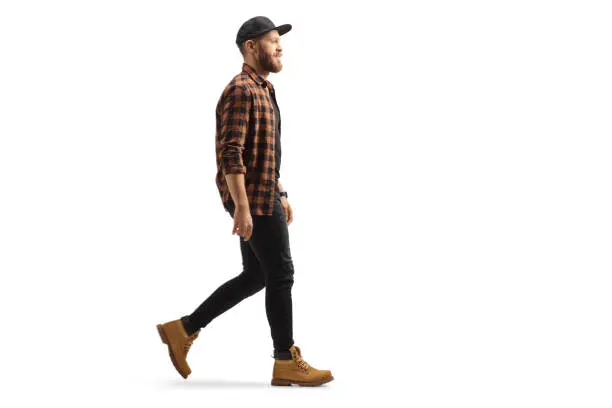 Full length profile shot of a young trendy man in a shirt and cap walking isolated on white background