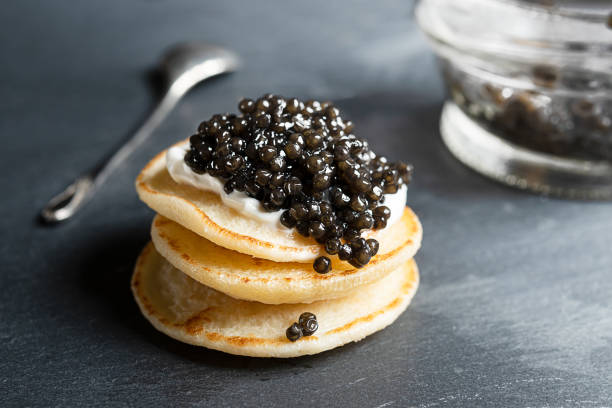 Black caviar on small pancakes Black sturgeon caviar on small blini pancakes, a glass jar and a spoon on a dark gray background caviar stock pictures, royalty-free photos & images