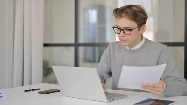 istock Young Man with Laptop Reading Documents in Modern Office 1363984034