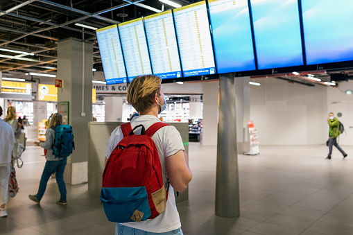 A young backpacker looking looking at departure/arrival screens at an airport. He is wearing a protective face mask to reduce the spread of COVID-19.
