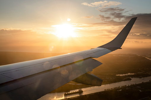 An aircraft wing on an airplane, flying above land mid flight while the sun is setting in the background.