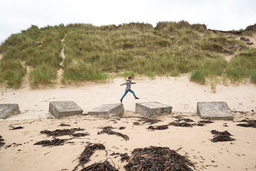 Wide shot view of a young boy playing at the beach in the North East of England. He is wearing warm clothing, running, and jumping across large rocks on the beach.