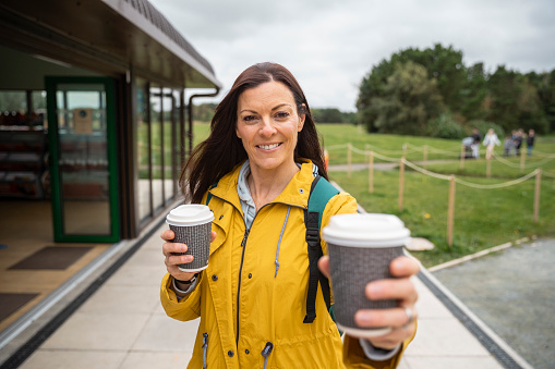A point of view shot of a mid-adult woman leaving a coffee shop with two cups of coffee, she is wearing warm clothing and passing a cup to her friend with a smile.