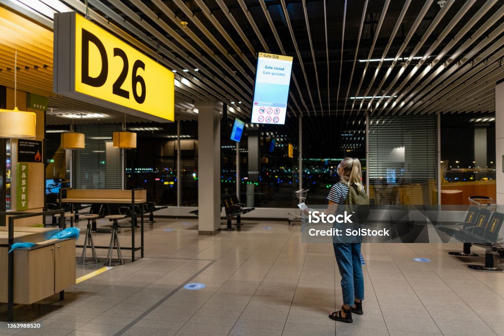 Catching a Connecting Flight A young backpacker looking looking at departure/arrival screens at an airport and her phone. She is wearing a protective face mask to reduce the spread of COVID-19. Airport Terminal Stock Photo