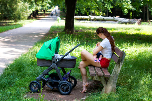 授乳中の女性は、ベビーカーの隣の公共公園の木製のベンチに座っています - female nurse 写真 ストックフォトと画像