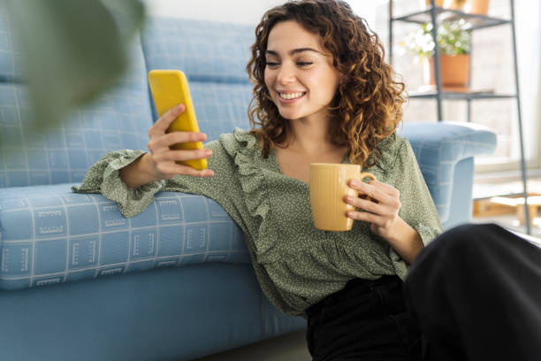mujer de pelo rizado sentada en el sofá con una taza de bebida caliente y teléfono móvil - mujeres jóvenes fotografías e imágenes de stock
