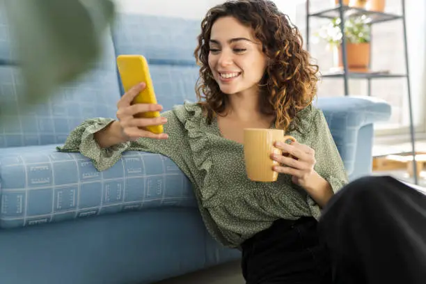 Photo of curly haired woman sitting on sofa with a cup of hot drink and mobile phone