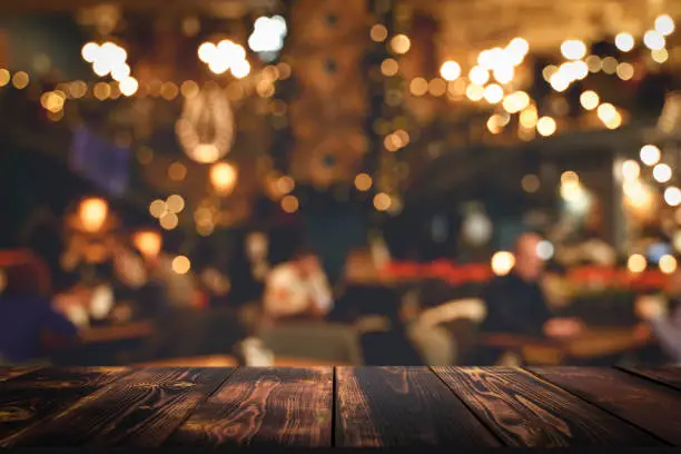 Photo of Table restaurant restaurant. Wooden kitchen or bar table on blurred interior of the evening restaurant hall with lights and visitors.