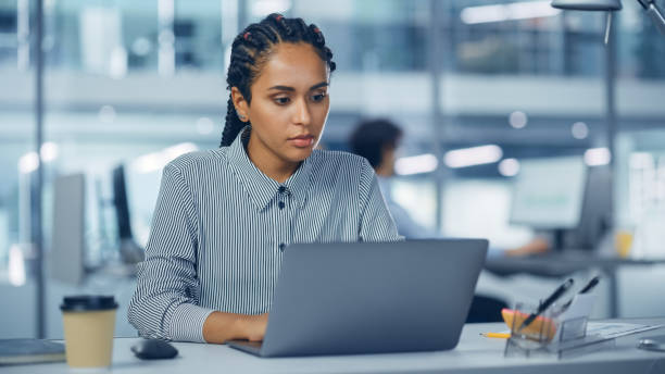 istock Multi-Ethnic Office: Professional Black Female IT Engineer Working on Desktop on Laptop Computer. Successful, Smiling Businesswoman with Braided Hair Does Creative Job. Confident Product Designer 1364004761